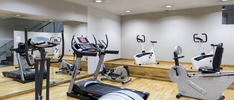 Un gimnasio con diferentes máquinas de cardio, piso de madera y un espejo grande.