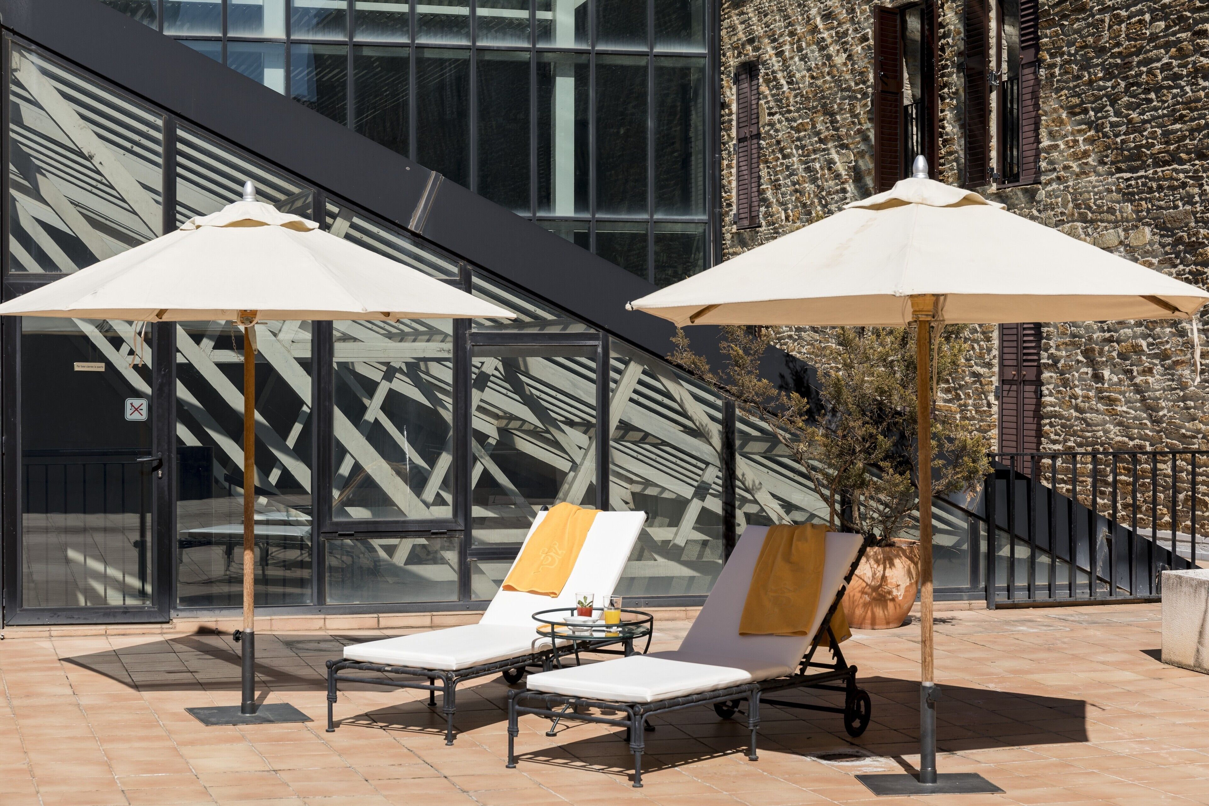 Two sun loungers with umbrellas, a glass wall, and a stone building in the background.