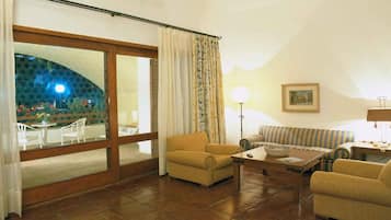 A living room with a sofa, armchairs, a coffee table, and a lamp. There is a large window with curtains, a painting on the wall, and a view of an outdoor area with tables and chairs.