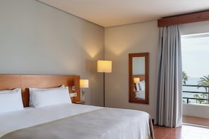 Una camera d'albergo con un letto grande, un comodino con una lampada, uno specchio e vista sul mare.
