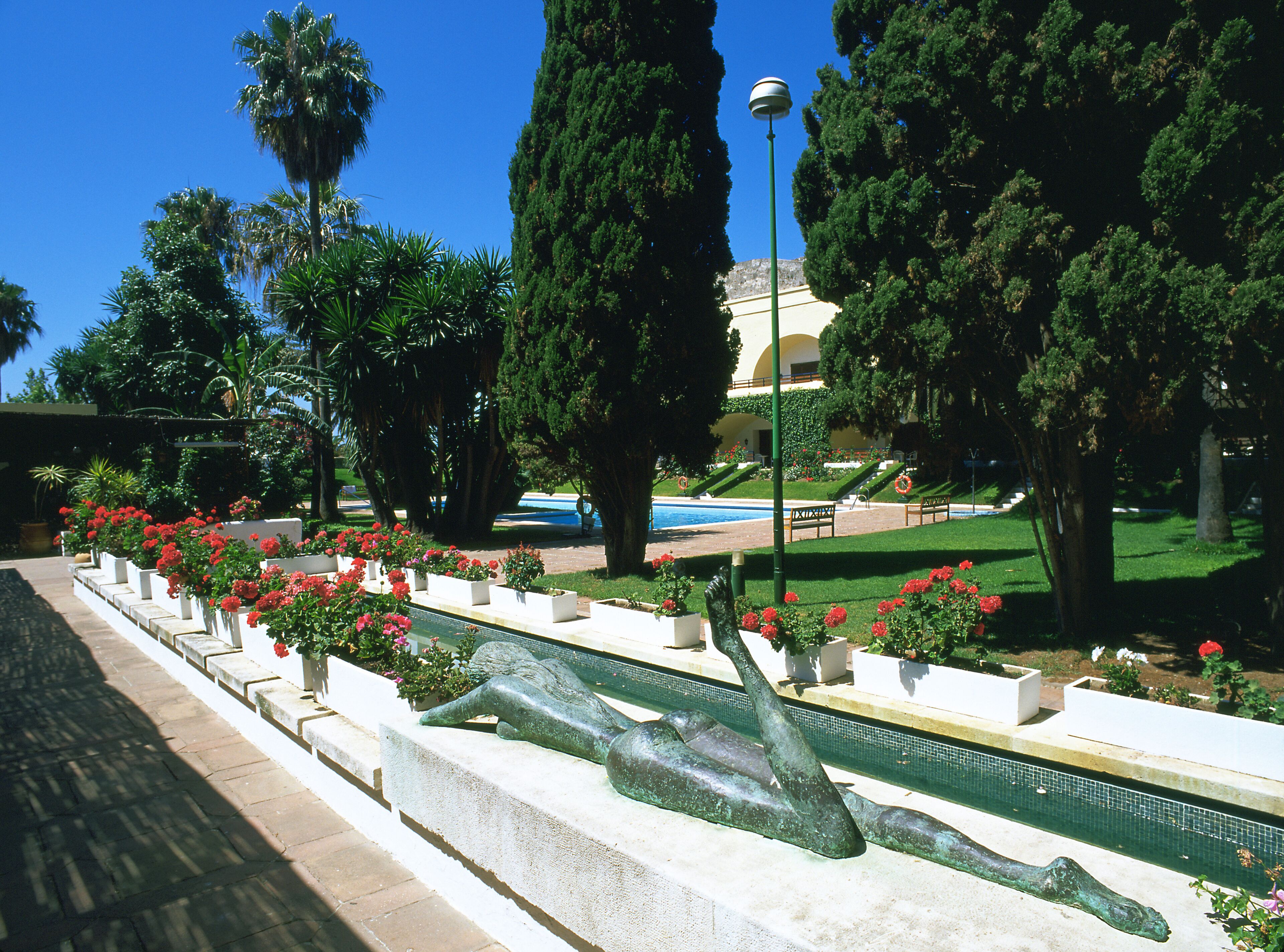 Un jardín ajardinado con piscina, bancos y una estatua.