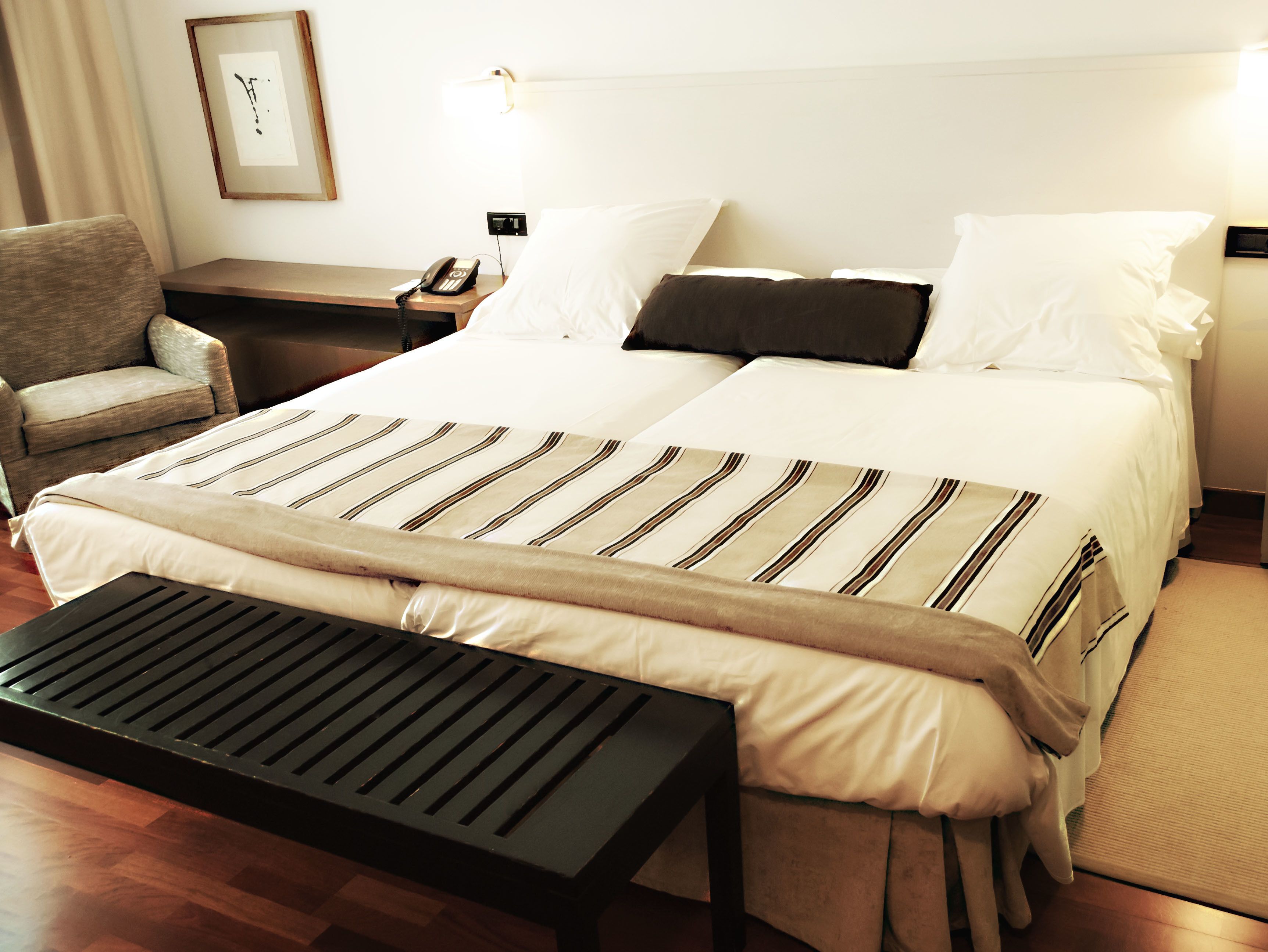 A hotel room with a large bed, a bedside table with a phone, a chair, and a bench.