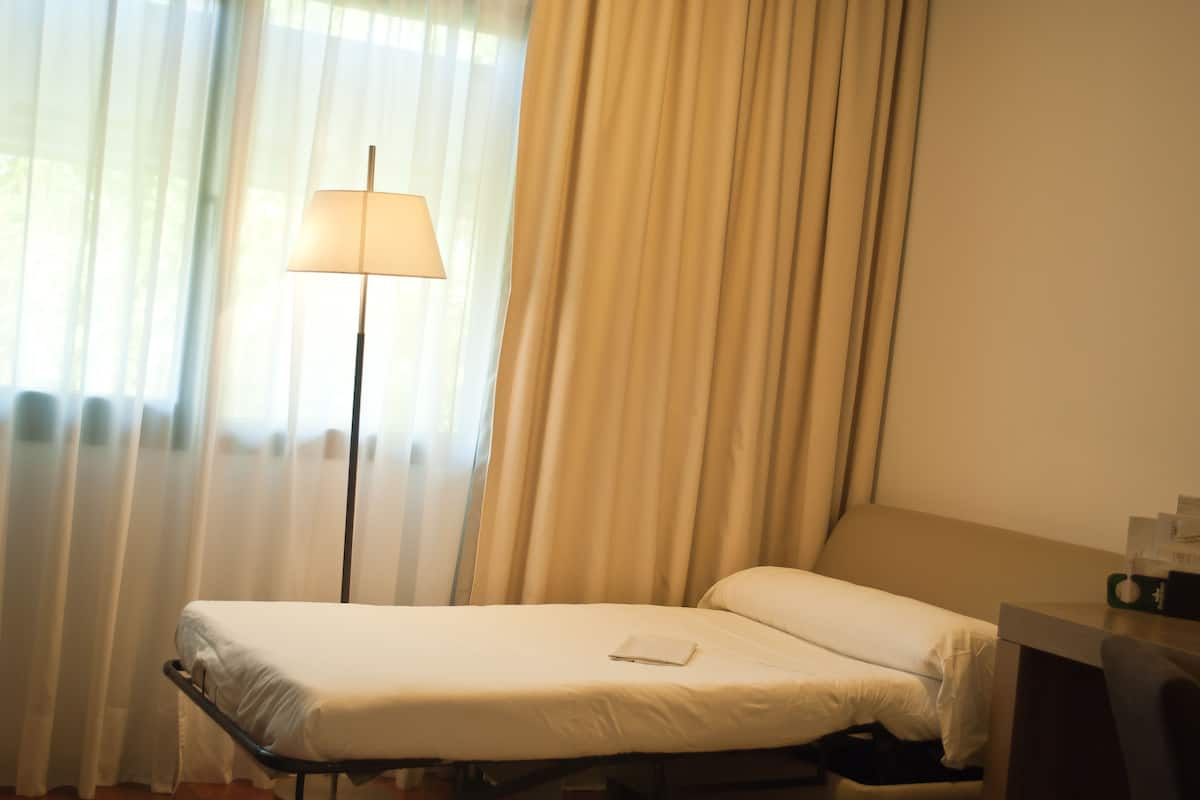 Un lit simple avec un matelas et des oreillers blancs, une table de chevet avec une lampe, et une fenêtre avec des rideaux légers.