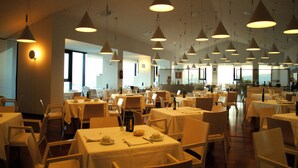 Een restaurant met meerdere tafels en stoelen, hangende verlichting aan het plafond en grote ramen voor natuurlijk licht.