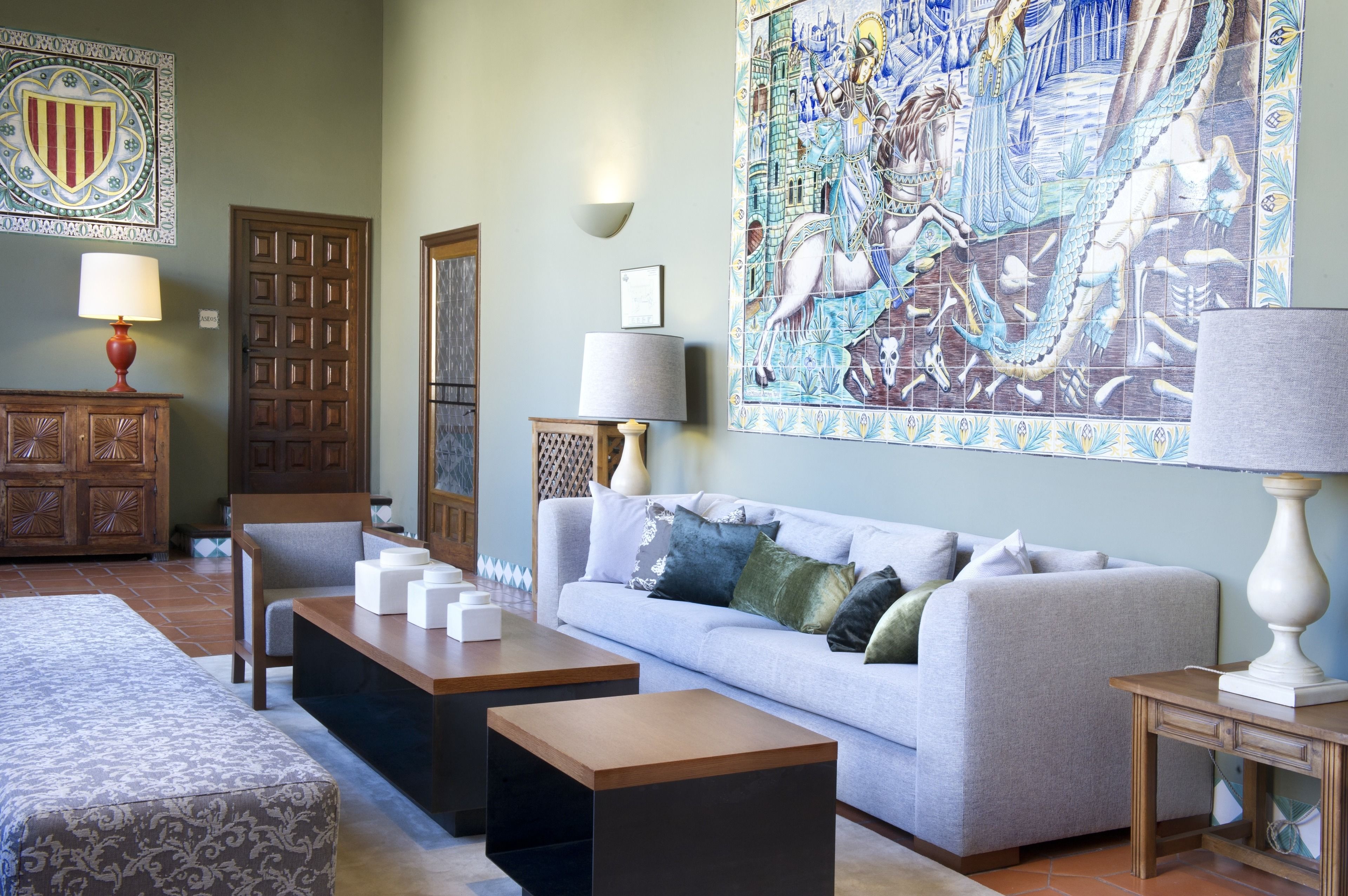 A living room with a light grey sofa, two wooden side tables, a patterned ottoman, and a large decorative tile mural on the wall.
