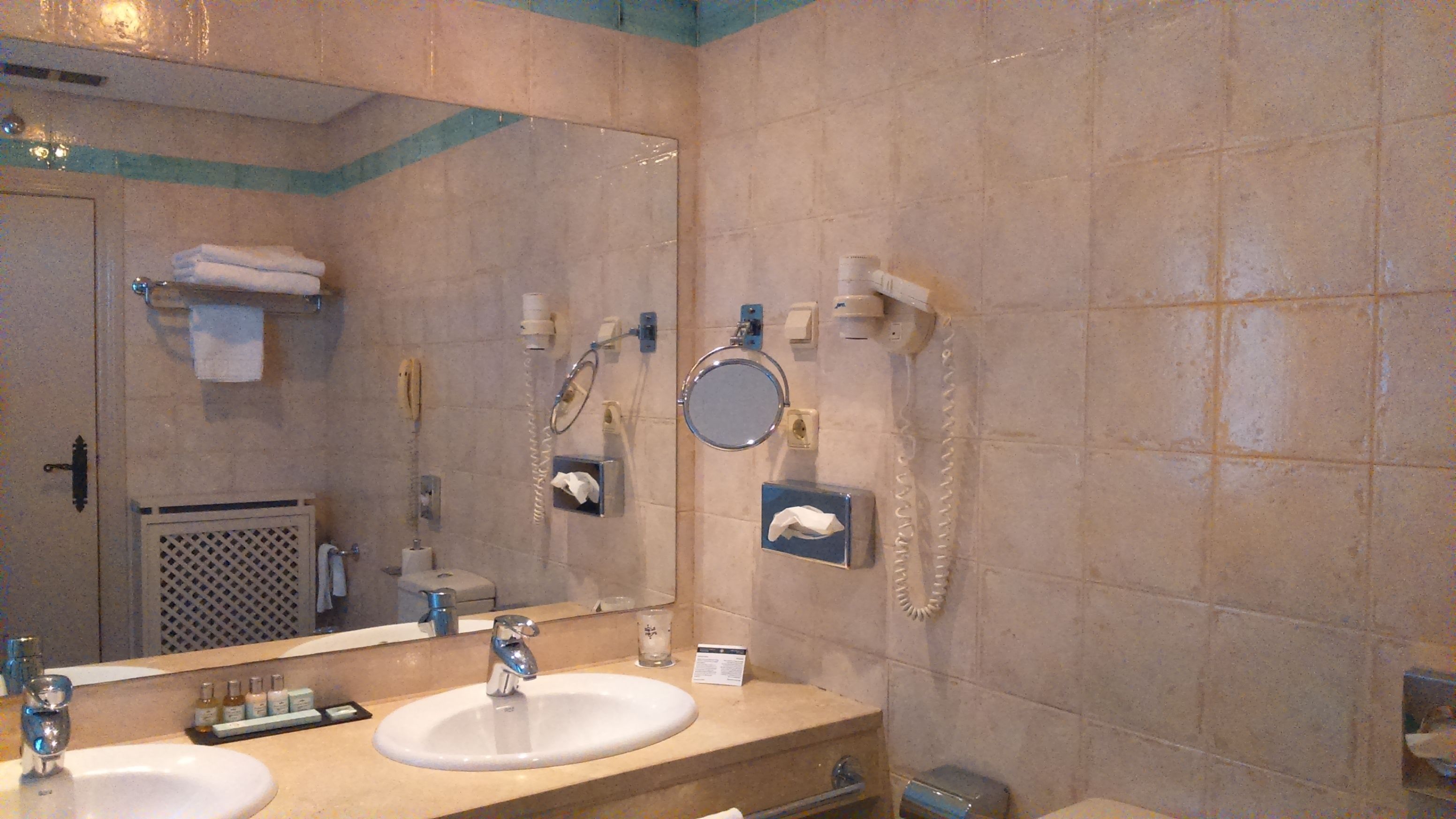 Un baño con dos lavamanos, un espejo grande, un perchero para toallas, un dispensador de papel higiénico y un secador de manos.