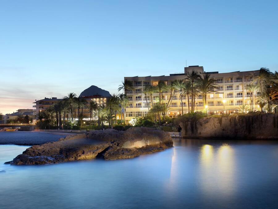 A hotel with multiple floors, palm trees, and a rocky shoreline.