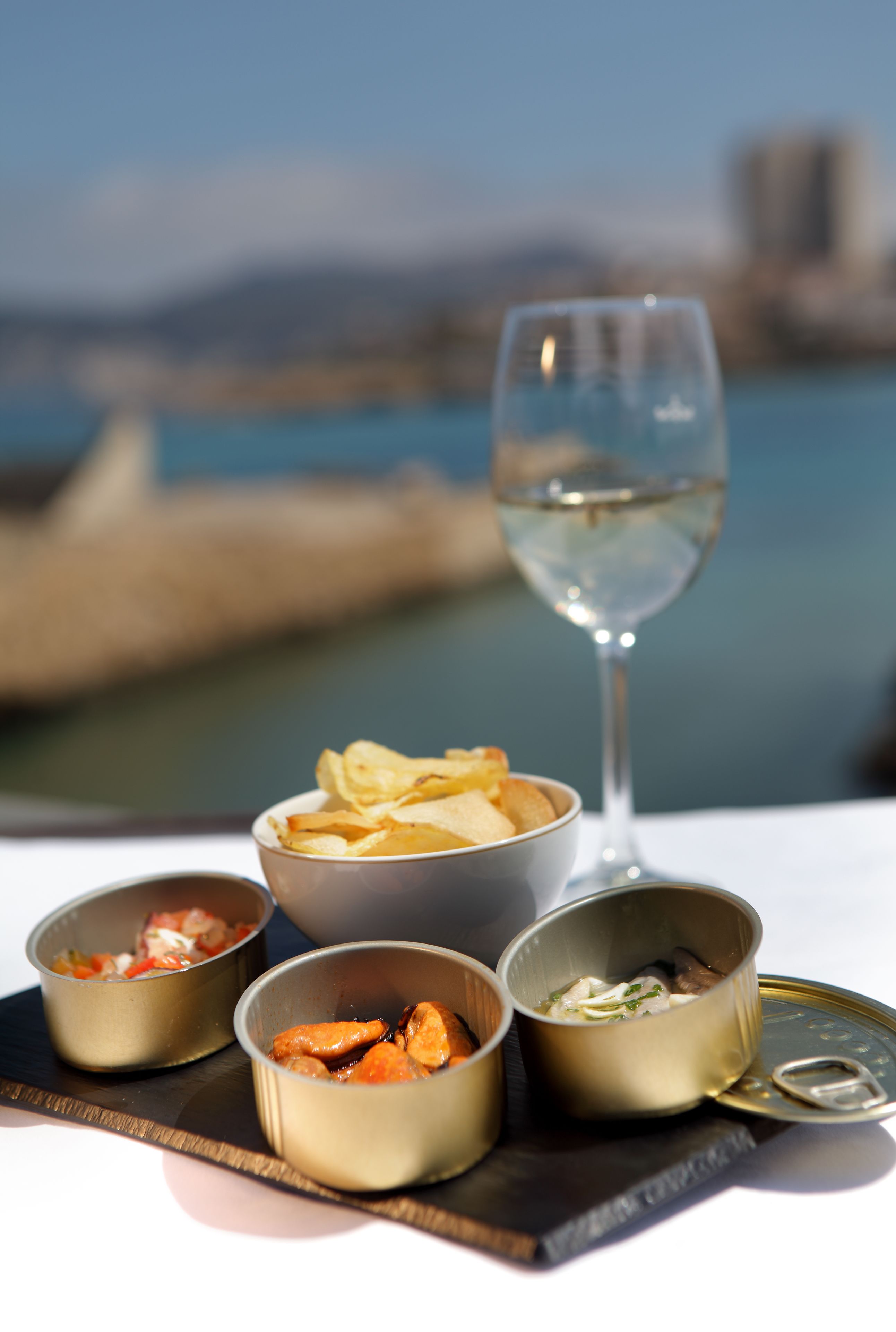 A table with four bowls of food and a glass of white wine, set against a backdrop of a body of water and distant buildings.
