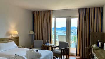 Habitación de hotel con cama, dos sillas, una mesita, escritorio y vistas al mar y a las montañas.