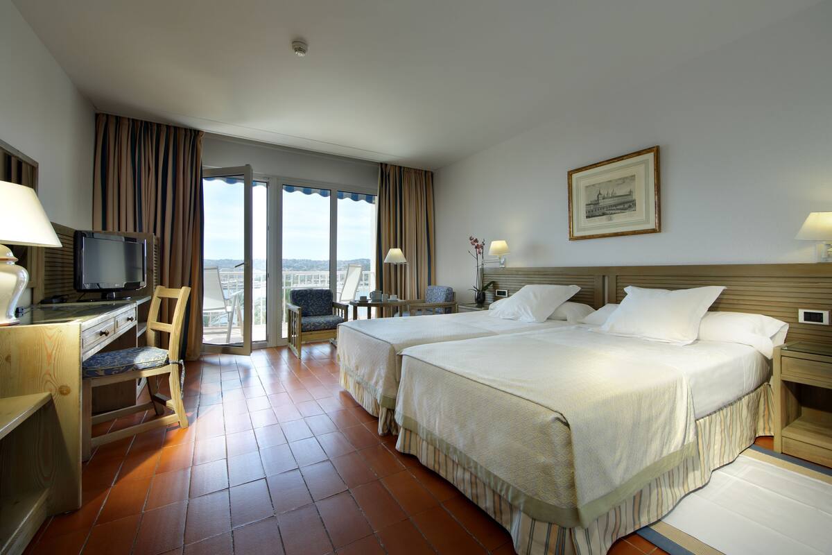 A hotel room with two beds, a desk with a television, a chair, and a view of the city.
