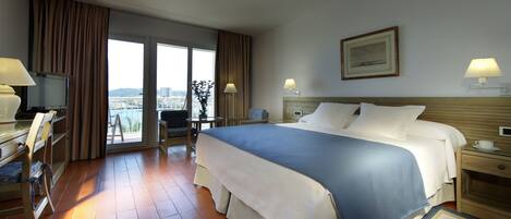 A hotel room with a large bed, a desk with a chair, a television, and a view of the city through the window.