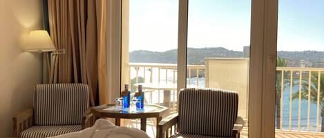 A hotel room with a bed, two striped armchairs, a small table with drinks, a lamp, and a balcony with a view of the sea.