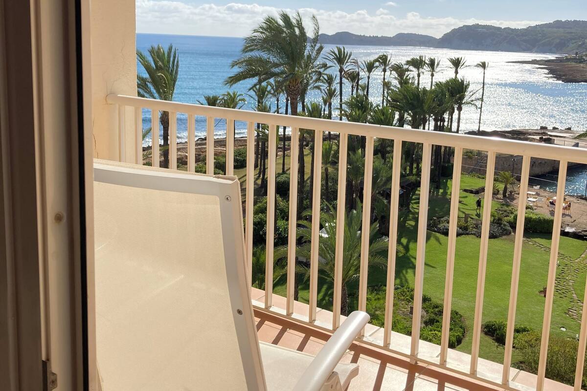 A balcony with a view of the sea, palm trees, and a grassy area.