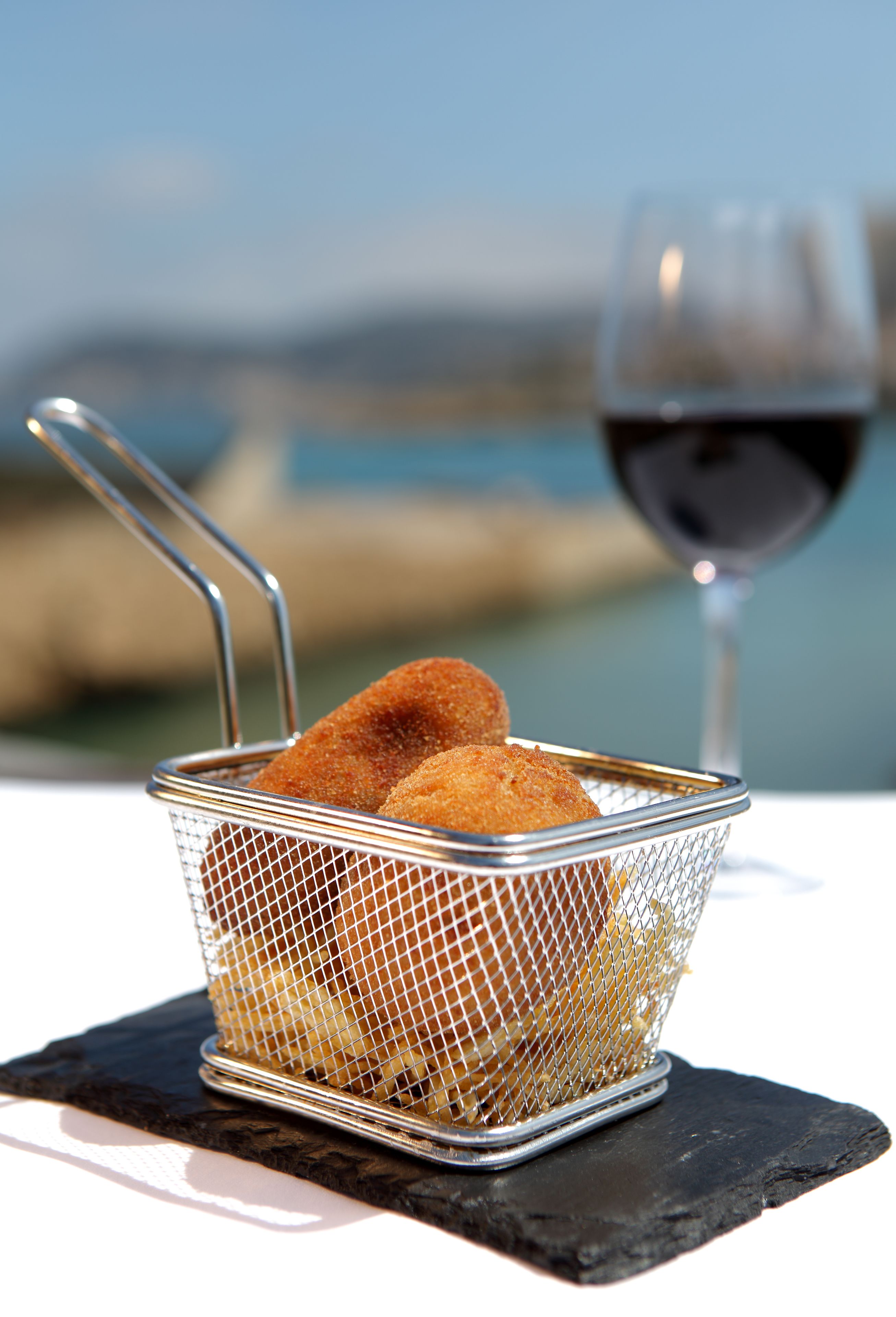 Una cesta de comida frita sobre un posavasos de pizarra, junto a una copa de vino tinto, con una vista panorámica de fondo.