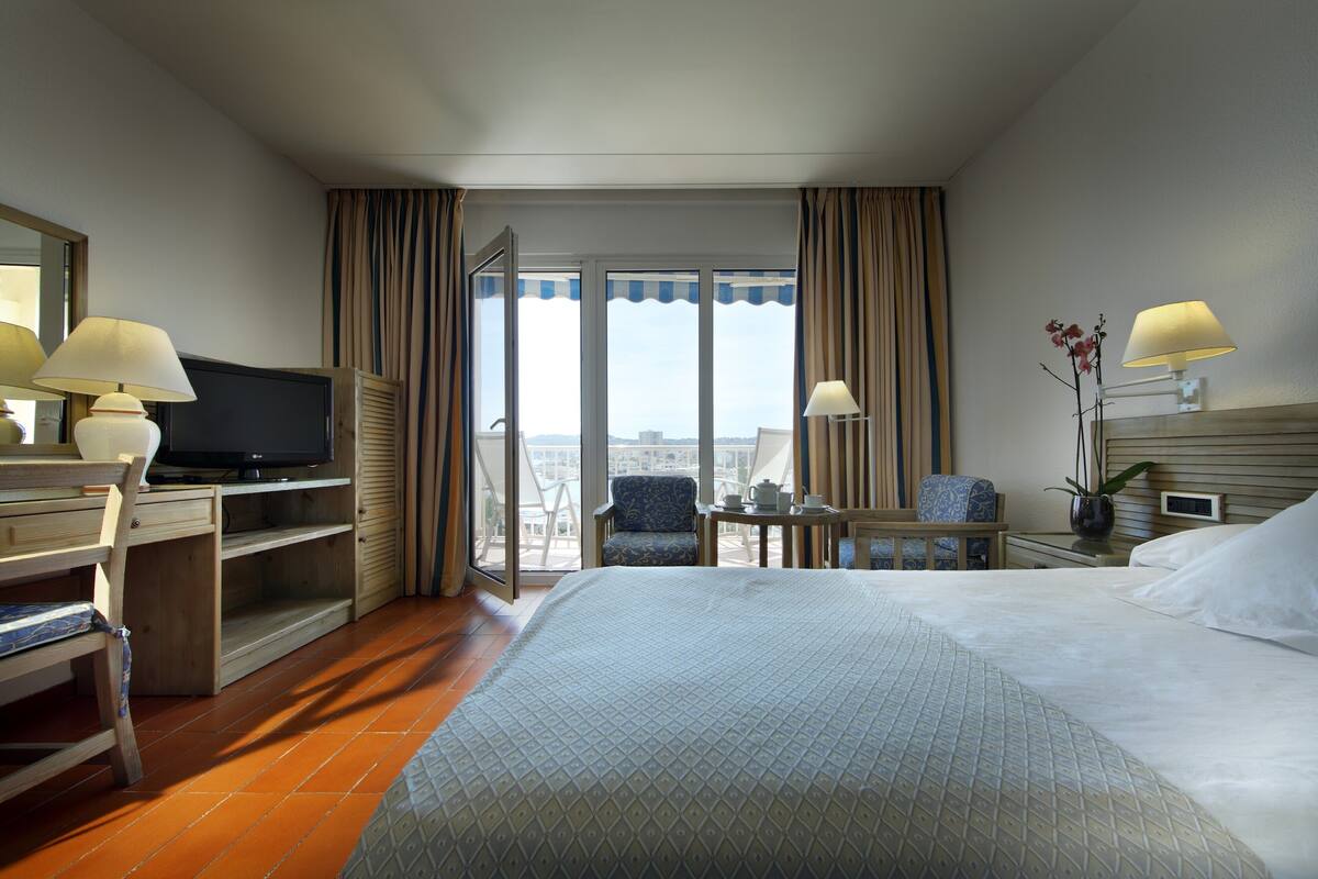A hotel room with a bed, a desk with a television, a chair, a small table with a lamp, and a view of the city through the window.