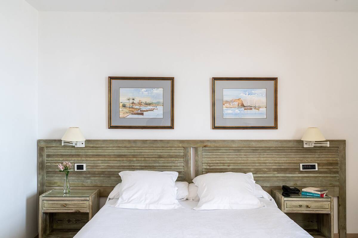 A hotel room with a wooden headboard, two bedside tables with lamps, a bed with white linens, and two framed paintings on the wall.