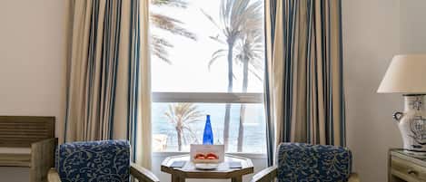 Una habitación con dos sillas azules con estampado, una pequeña mesa de madera con una botella y fruta, y vistas a palmeras y al mar a través de cortinas a rayas.