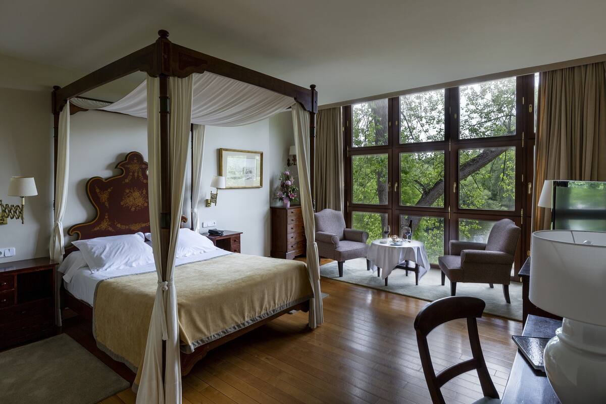 Una cama con dosel, un ventanal con vista a árboles, una mesita redonda y dos sillones.