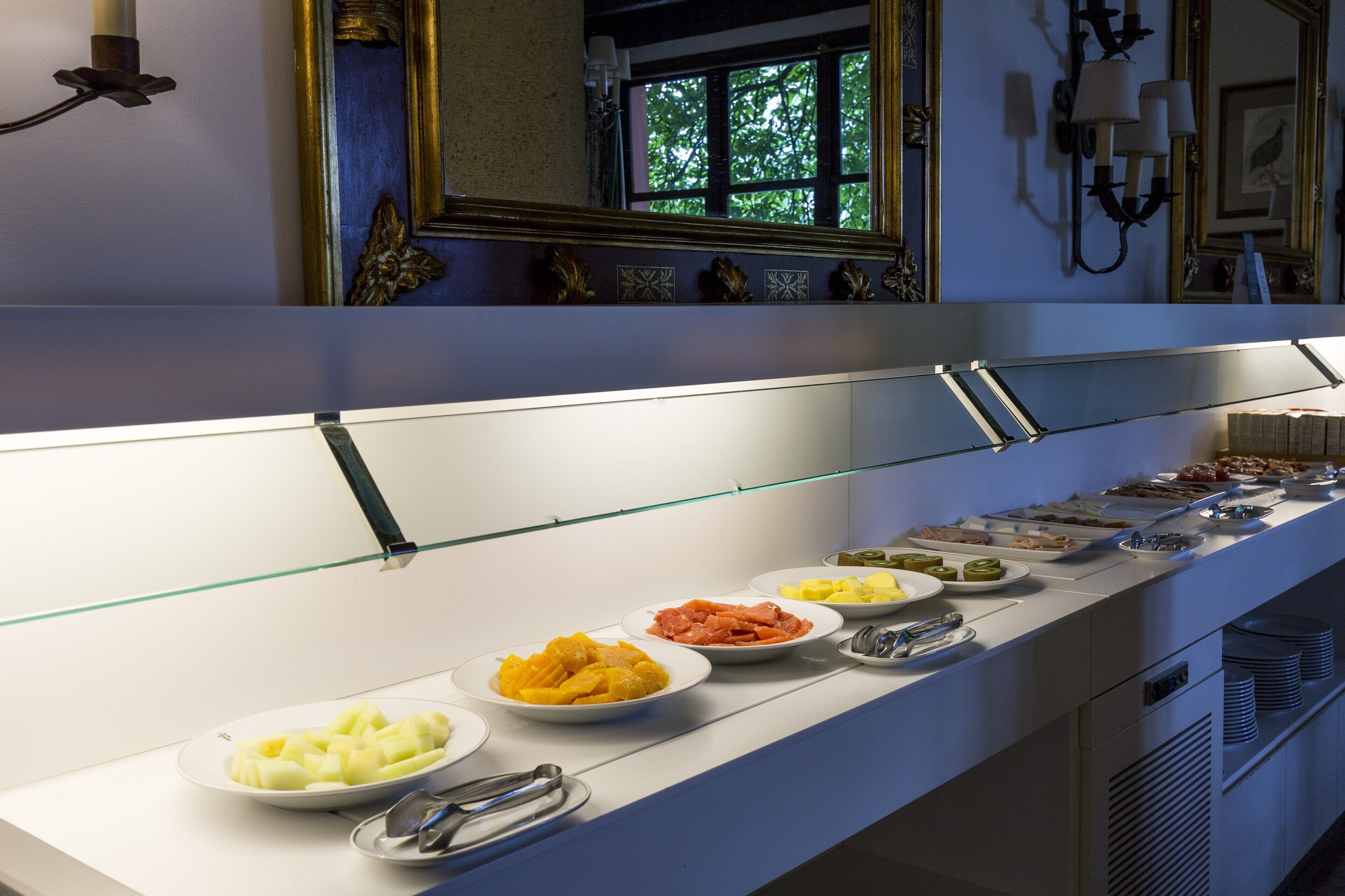 Un bufé con diversos alimentos, entre ellos frutas y carnes, en una habitación bien iluminada con ventana y apliques decorativos.