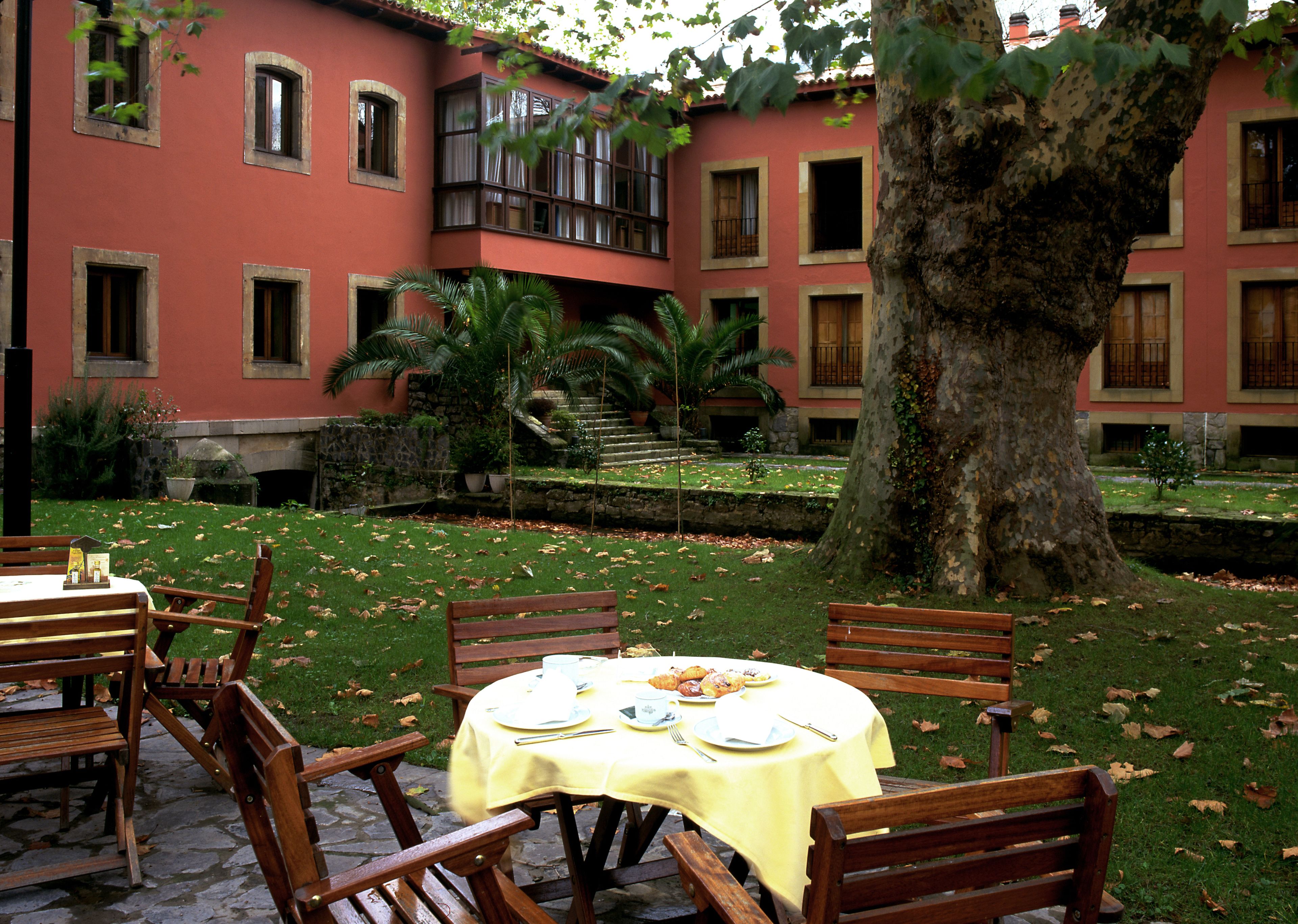 Un patio con una mesa dispuesta para una comida, rodeado de sillas de madera y un gran árbol en el centro.