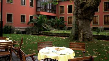 Un patio con una mesa dispuesta para una comida, rodeado de sillas de madera y un gran árbol en el centro.