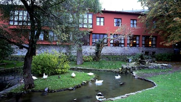 Un estanque con patos, rodeado de hierba y árboles, y un edificio rojo al fondo.