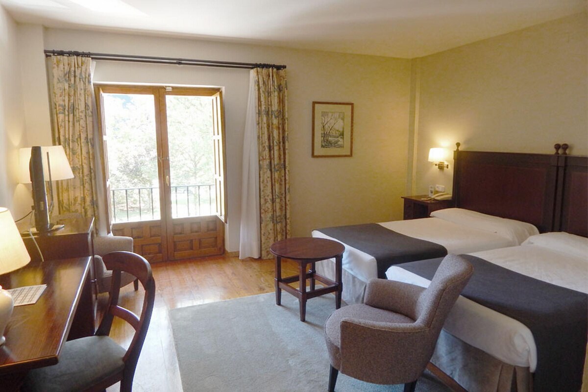 A hotel room with two beds, a desk with a chair, a small table, and a window with curtains.