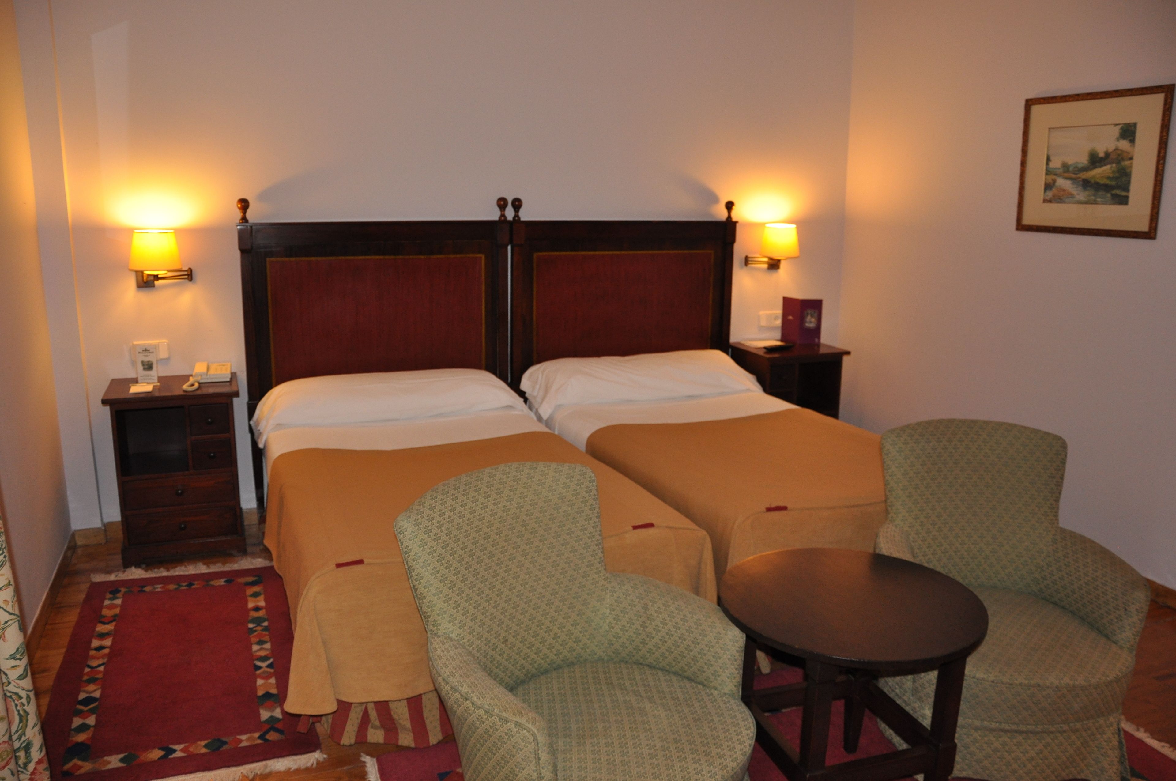 A hotel room with two beds, a nightstand, a chair, a small table, and two wall lamps.