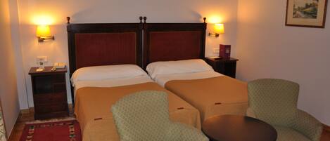A hotel room with two beds, a nightstand, a chair, a small table, and two wall lamps.