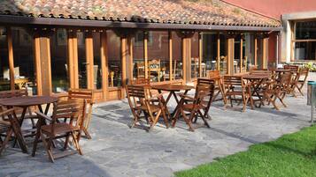 An outdoor patio with wooden tables and chairs, stone flooring, and a tiled roof.