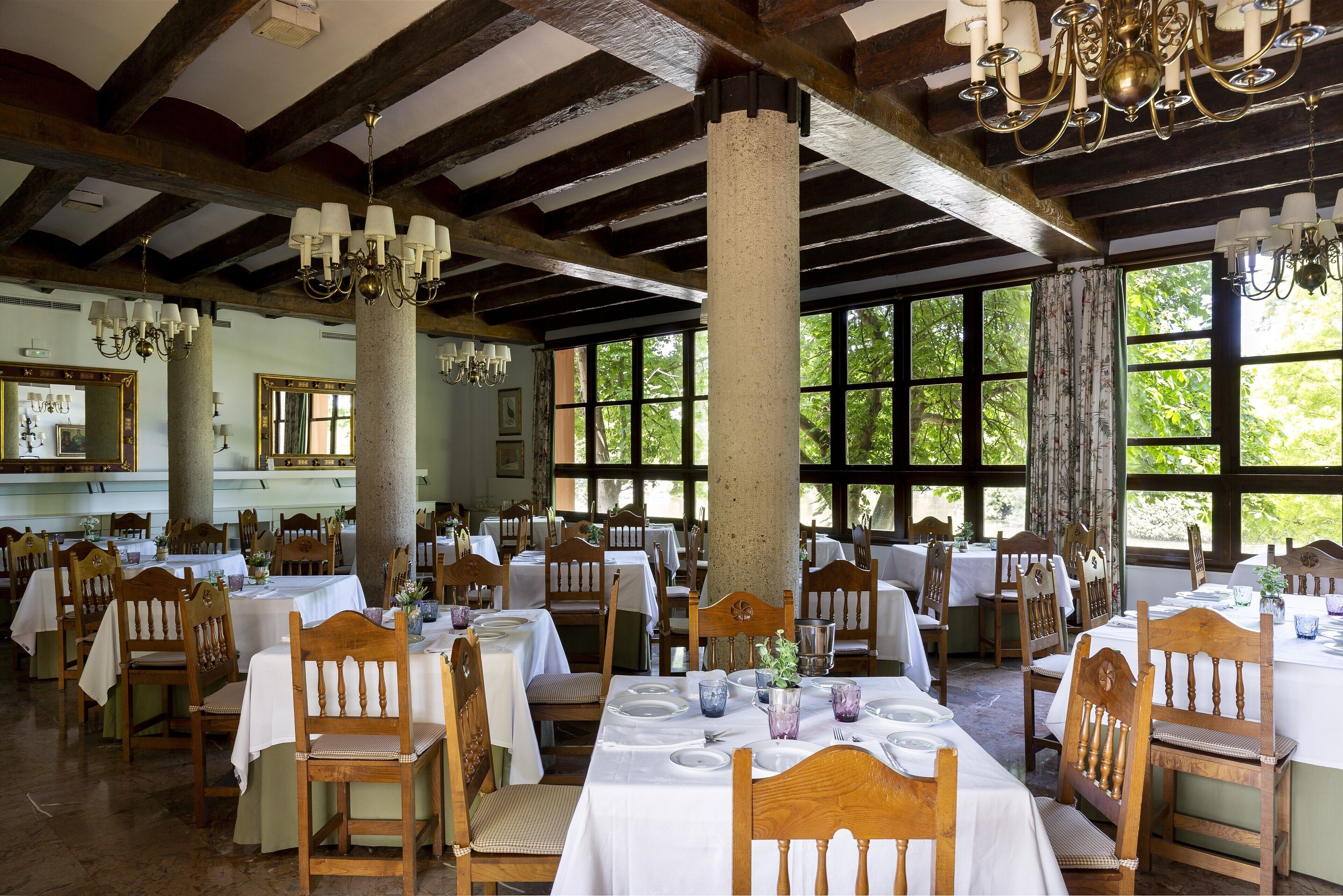 Un comedor con mesas y sillas de madera, ventanas amplias y candelabros.