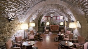 Un interior de paredes de piedra con techos abovedados, varias mesas y sillas, y lámparas fijadas a la pared.
