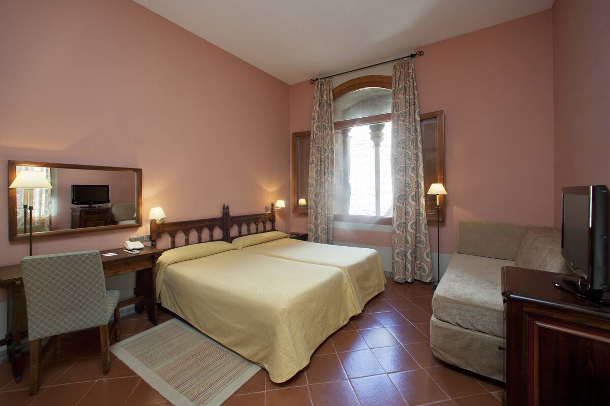 Habitación de hotel con dos camas, un escritorio, una silla, un televisor y una ventana con cortinas.