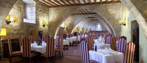 A restaurant with stone walls, wooden beams, and striped chairs. Tables are set with white tablecloths and wine glasses.