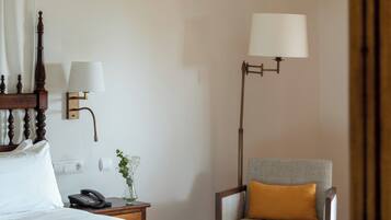 Habitación de hotel con cama, mesita de noche con lámpara, una silla con cojín y un espejo que refleja parte de la habitación.