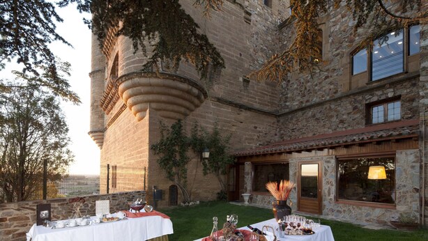 Mobiliario de comedor al aire libre con dos mesas, cada una con mantel blanco y borde rojo y verde, frente a un edificio de piedra con torre y hiedra.