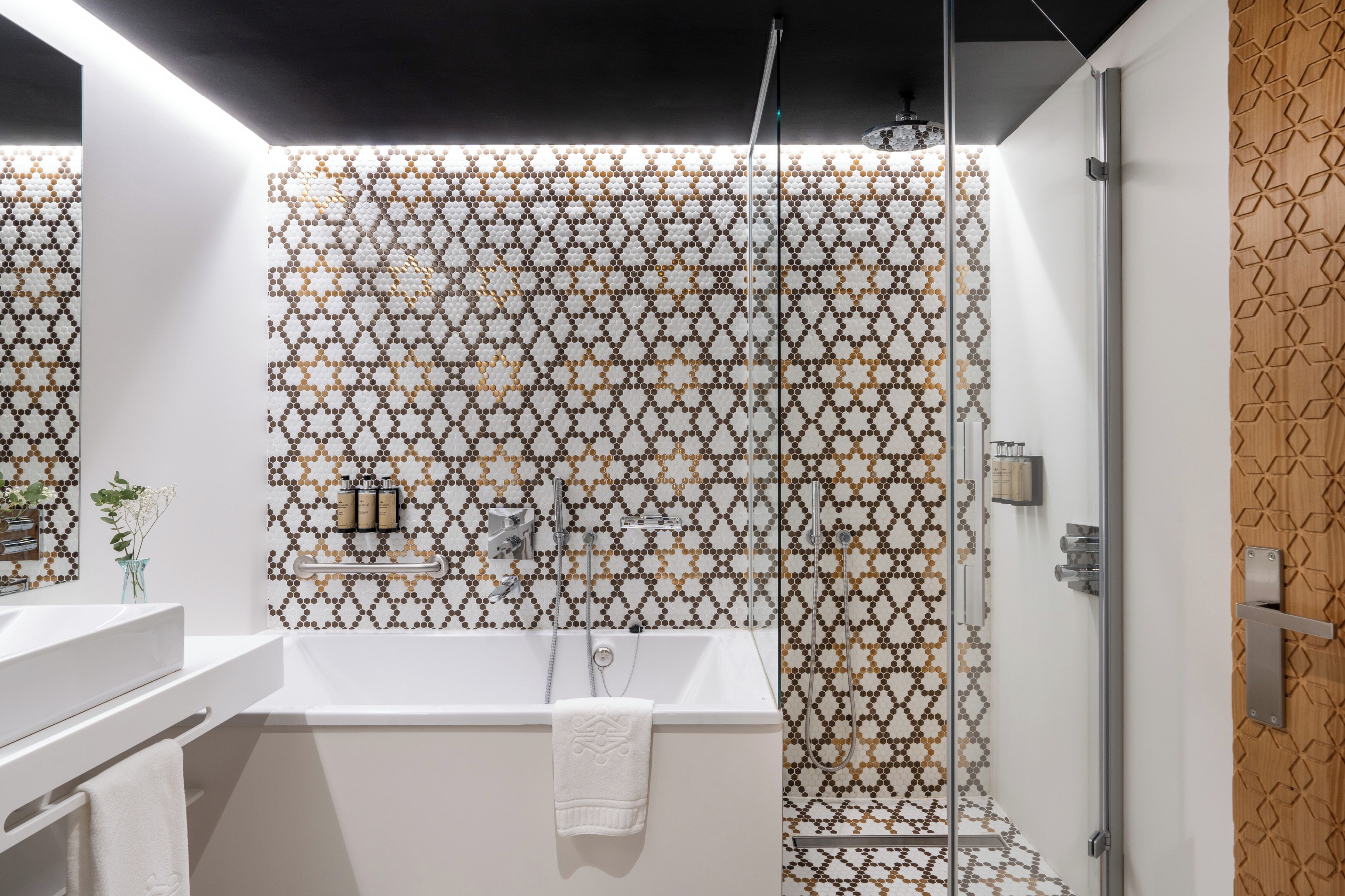 Um banheiro moderno com parede de azulejos estampados, banheira branca, box de vidro e porta de madeira.