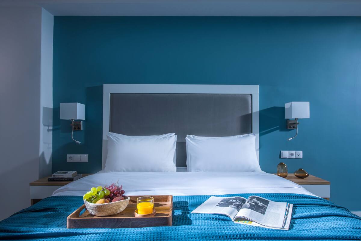 Ein ordentlich bezogenes Bett mit weißen Leinentüchern, einer blauen, strukturierten Decke, einem Tablett mit Früchten und Saft sowie einer Zeitschrift auf dem Bett.