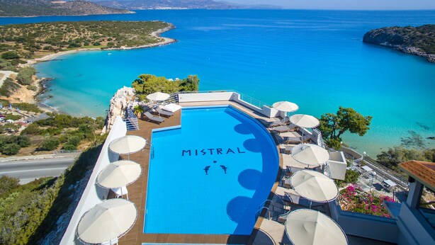Una piscina con la scritta "MISTRAL", circondata da sdraio e ombrelloni, affacciata su una zona costiera con acque blu cristalline e montagne in lontananza.