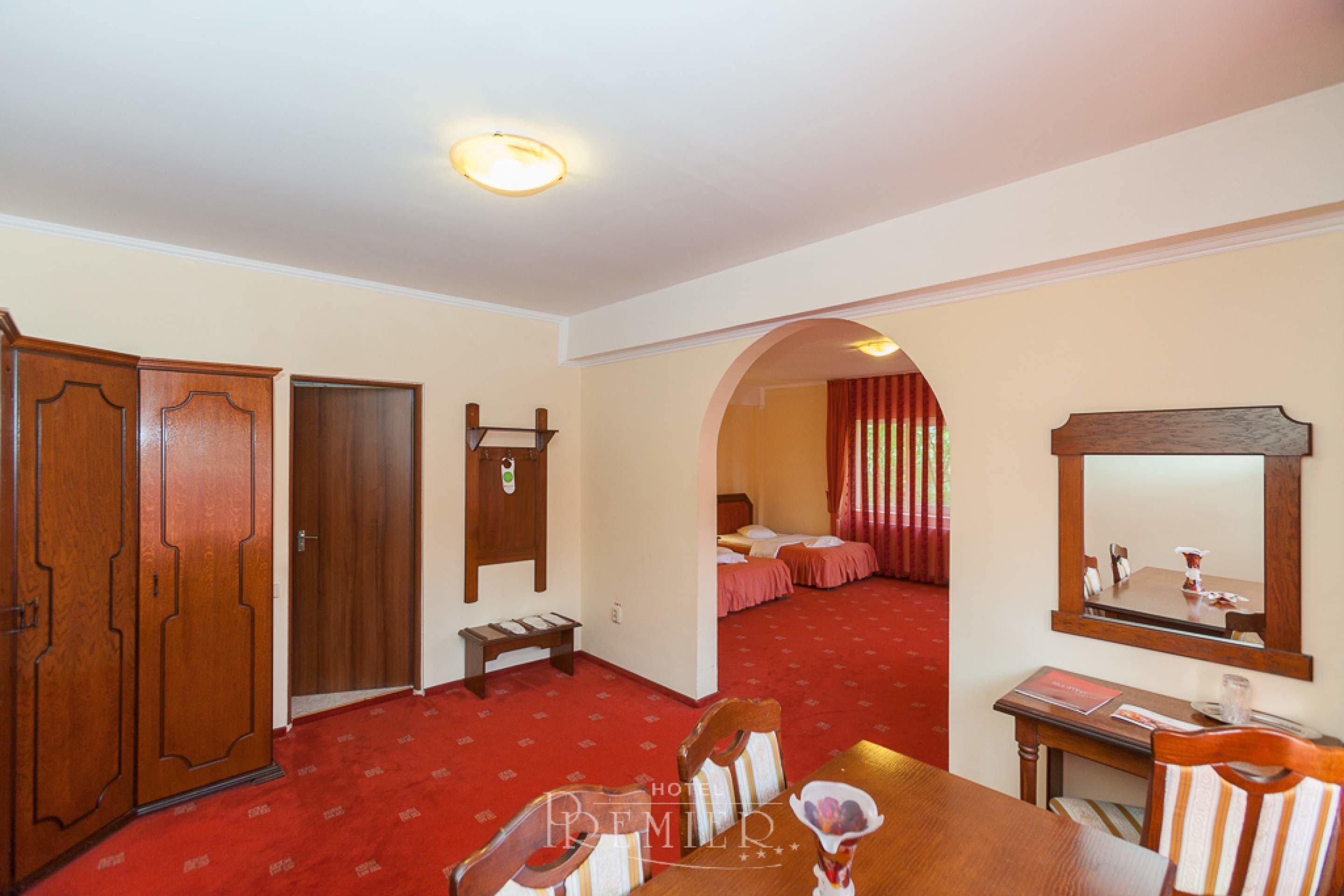 A hotel room with a red carpet, wooden wardrobe, a bed with pink curtains, a mirror, and a dining area with chairs and a table.