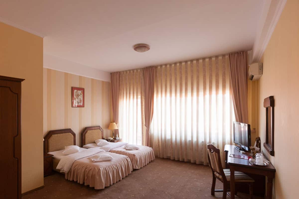 Ein Hotelzimmer mit zwei Betten, einem Schreibtisch mit Stuhl, einem Fernseher und einem Fenster mit Vorhängen.