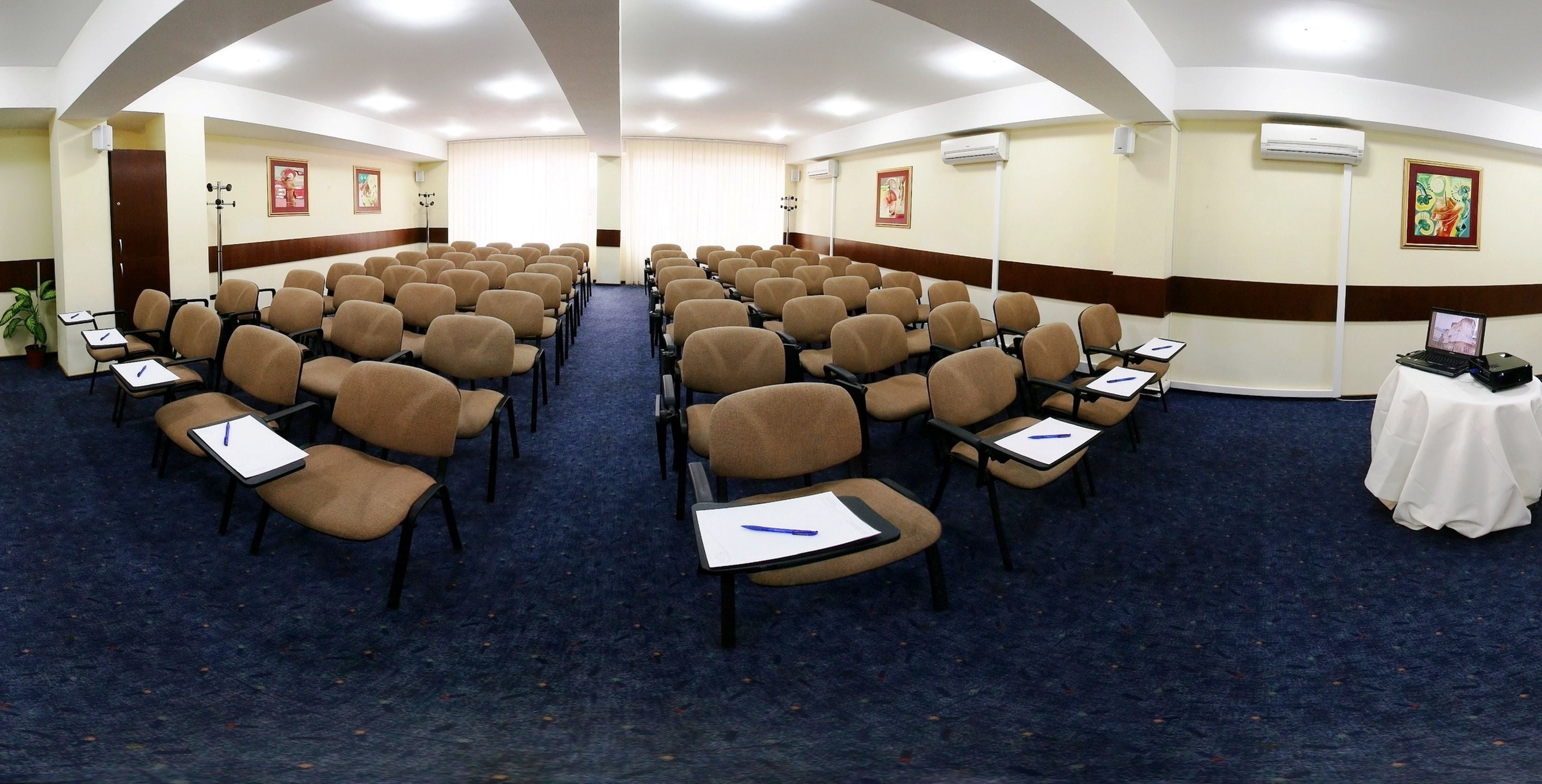 Un salón de conferencias con filas de sillas, mesas con blocs de notas y unidades de aire acondicionado.