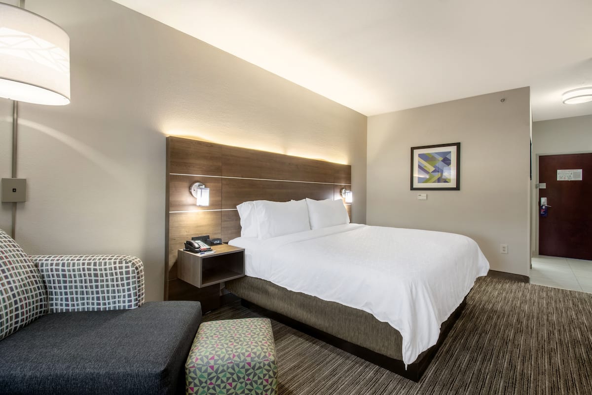 A hotel room with a large bed, a bedside table with a lamp, a chair, and a patterned ottoman.