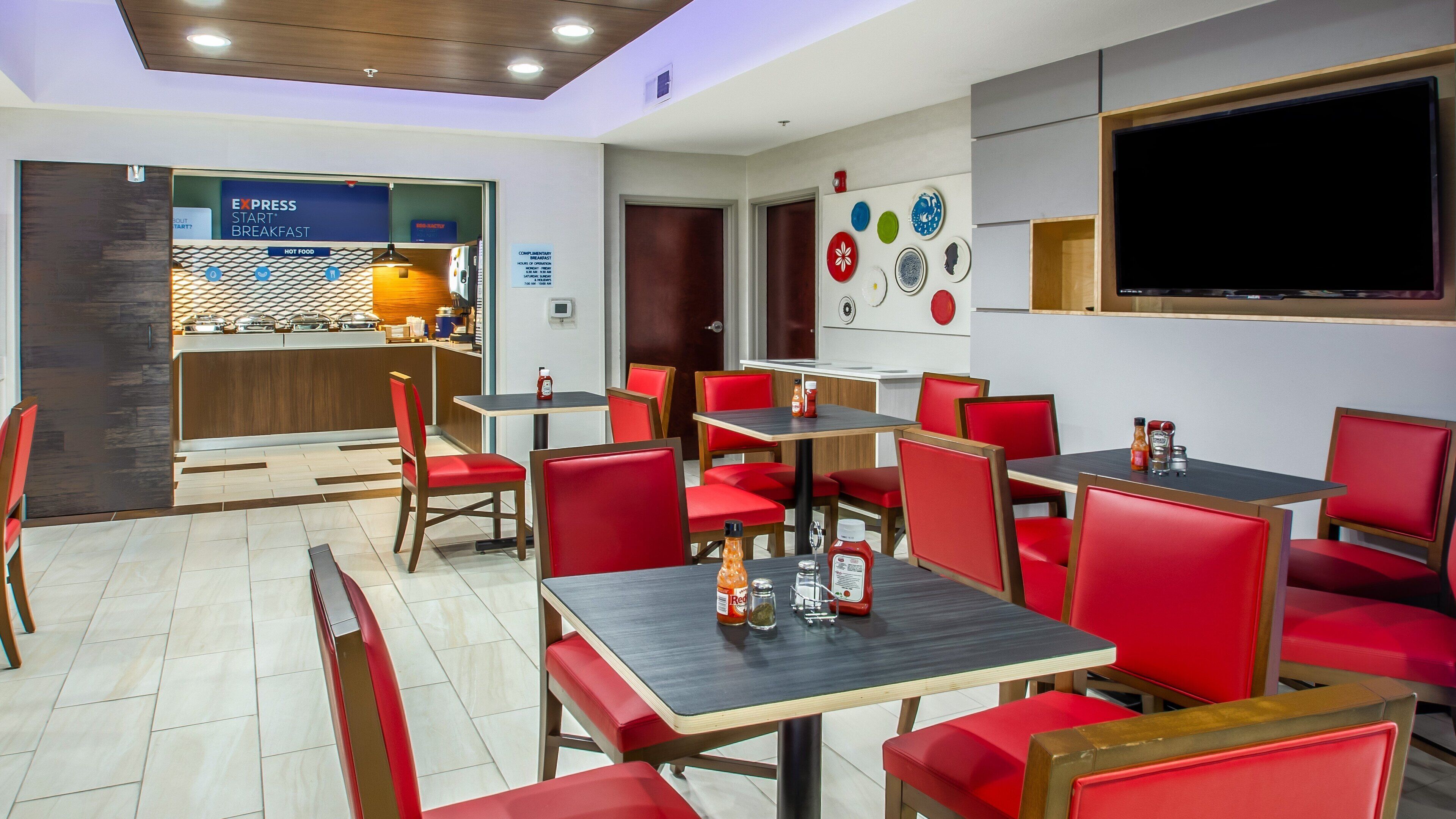 A restaurant with red upholstered chairs and tables, a kitchen area, and a flat-screen TV on the wall.