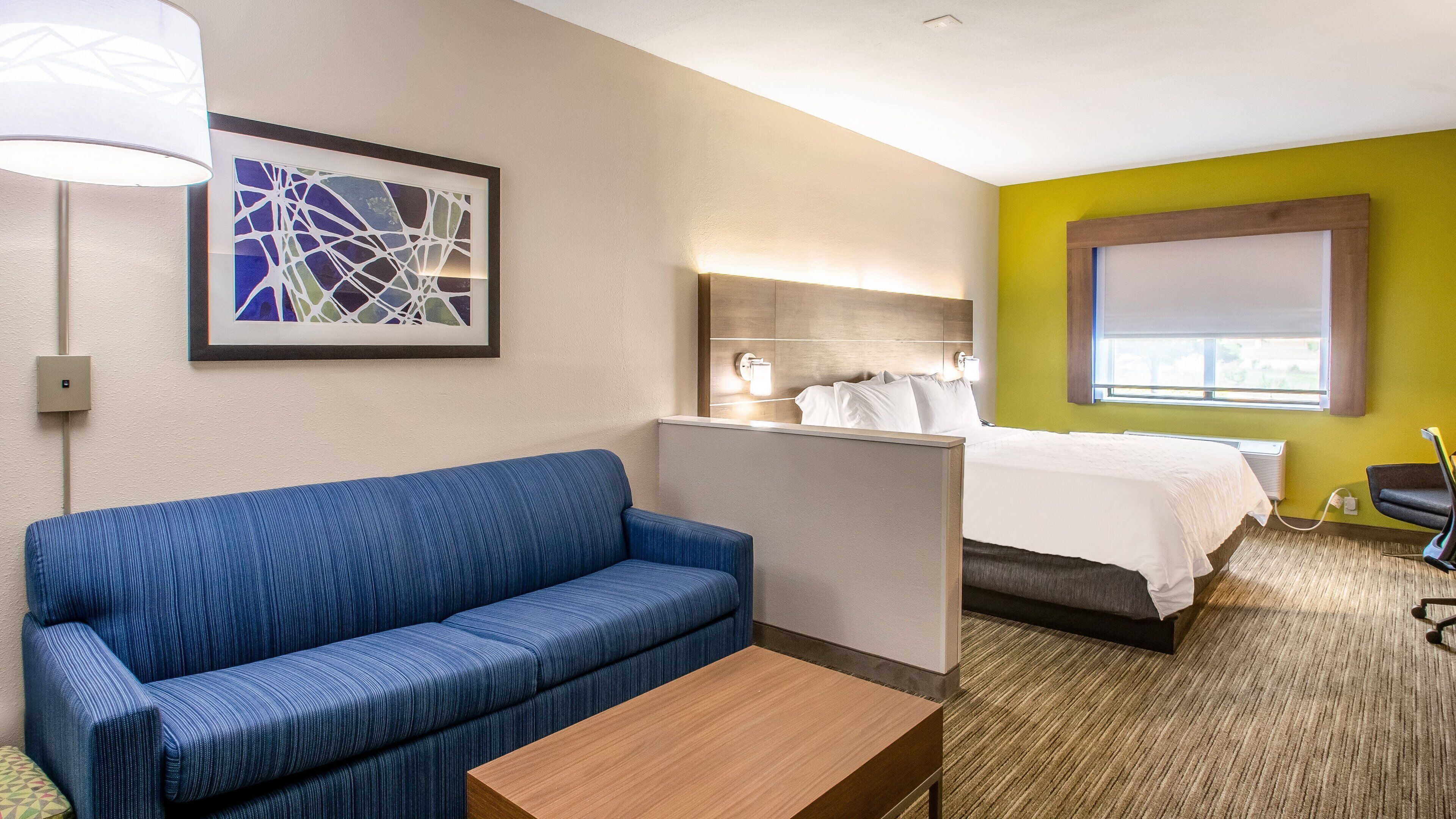 A hotel room with a blue sofa, a wooden coffee table, a bed with white bedding, a nightstand, a lamp, and a window with blinds.