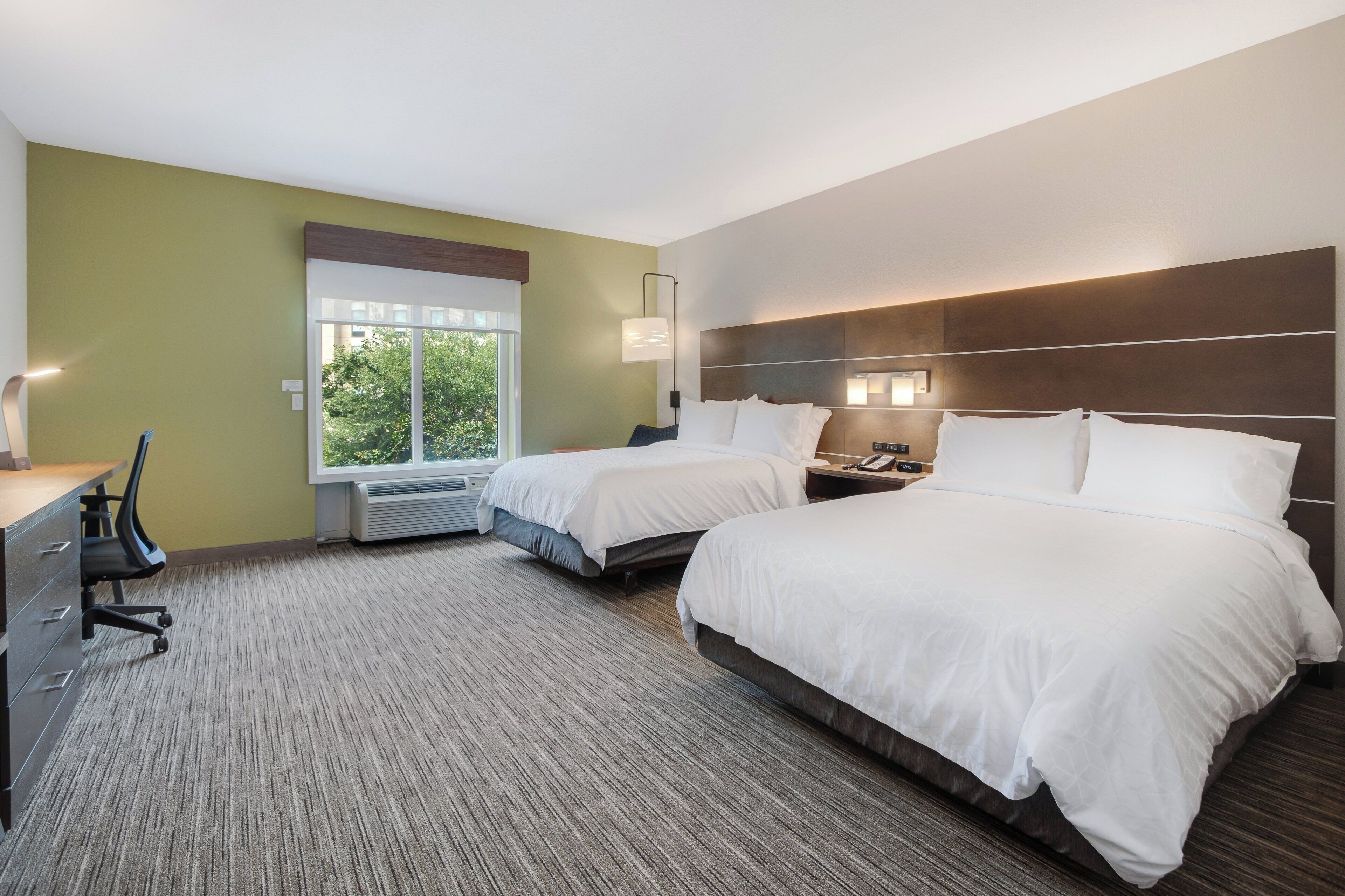 A hotel room with two beds, a desk, a chair, a window, and a wall-mounted lamp.
