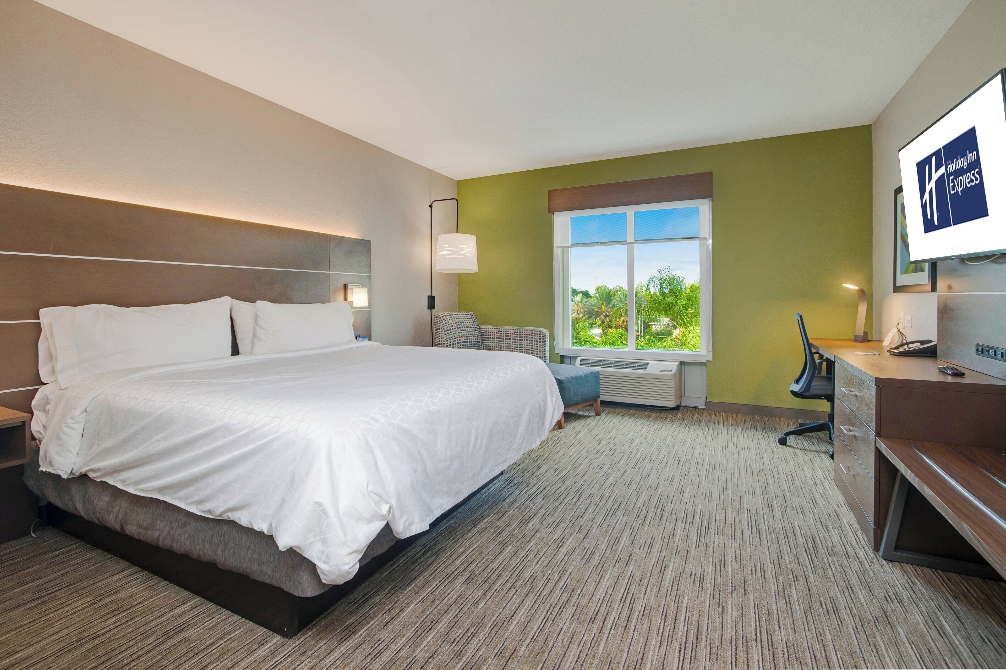 A hotel room with a large bed, a desk, a chair, a TV, and a window with a view of greenery.