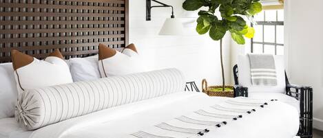A neatly arranged bedroom with a large bed, a headboard with a grid pattern, a bedside table with a lamp, a potted plant, and a chair with a towel.