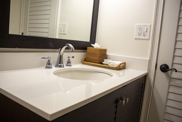 A bathroom with a white sink, two faucets, a mirror, a basket with a tissue, and a towel. - Inn On The Lakes (Sebring)