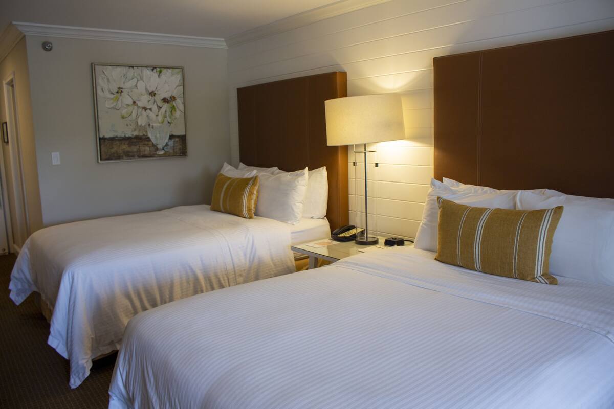 Quarto de hotel com duas camas, uma mesa de cabeceira com abajur, um quadro na parede e um telefone.